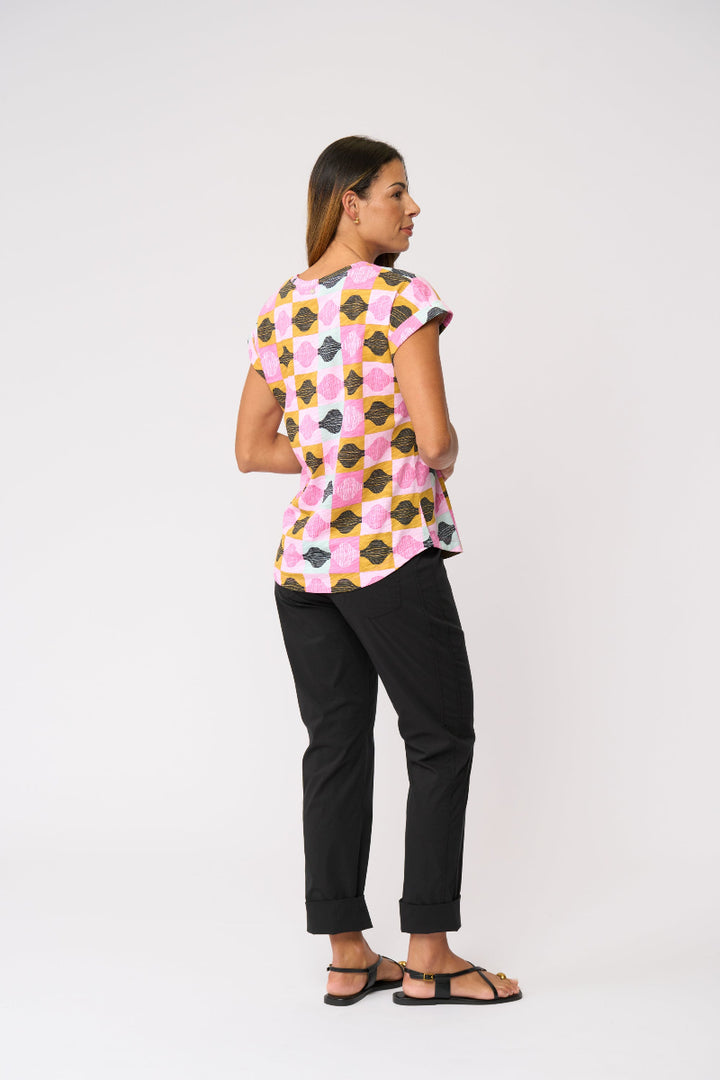 A woman is shown from the back wearing a short-sleeve top with a colorful geometric pattern of pink, yellow, black, and white shapes, paired with black pants and black sandals, against a plain white studio background.
