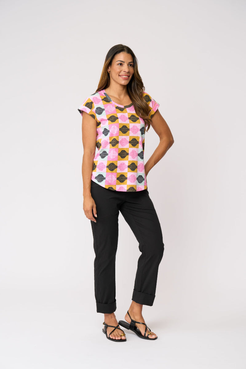 A woman modeling a short-sleeve crewneck top with a pink, yellow, and black geometric pattern, paired with black pants and sandals, against a plain light gray background.