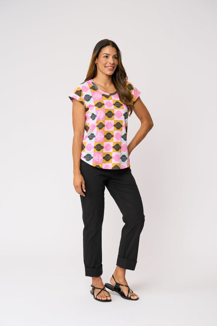 A woman modeling a short-sleeve crewneck top with a pink, yellow, and black geometric pattern, paired with black pants and sandals, against a plain light gray background.