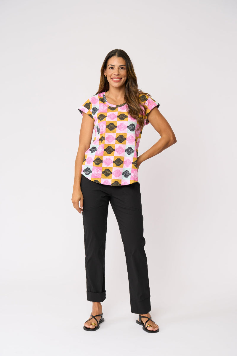 A woman modeling a short-sleeve crewneck top with a colorful geometric pattern in pink, yellow, black, and white, paired with black pants and sandals, against a plain white studio background.