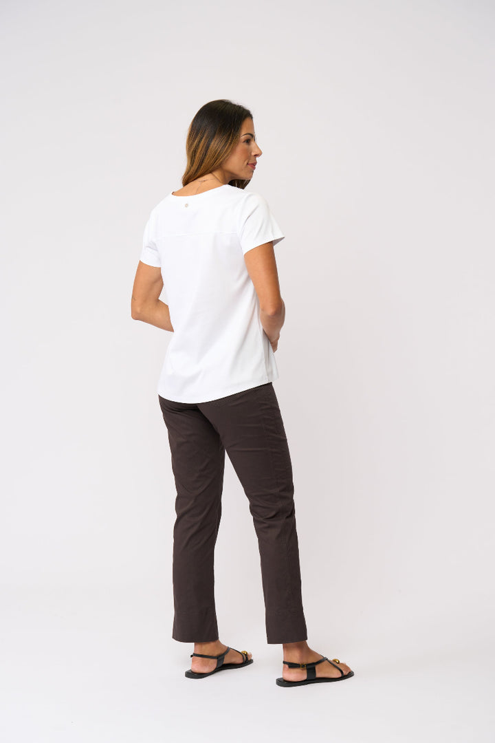 A woman is shown from the back wearing a white short-sleeve t-shirt, dark brown cropped pants, and black sandals against a plain light gray studio background.