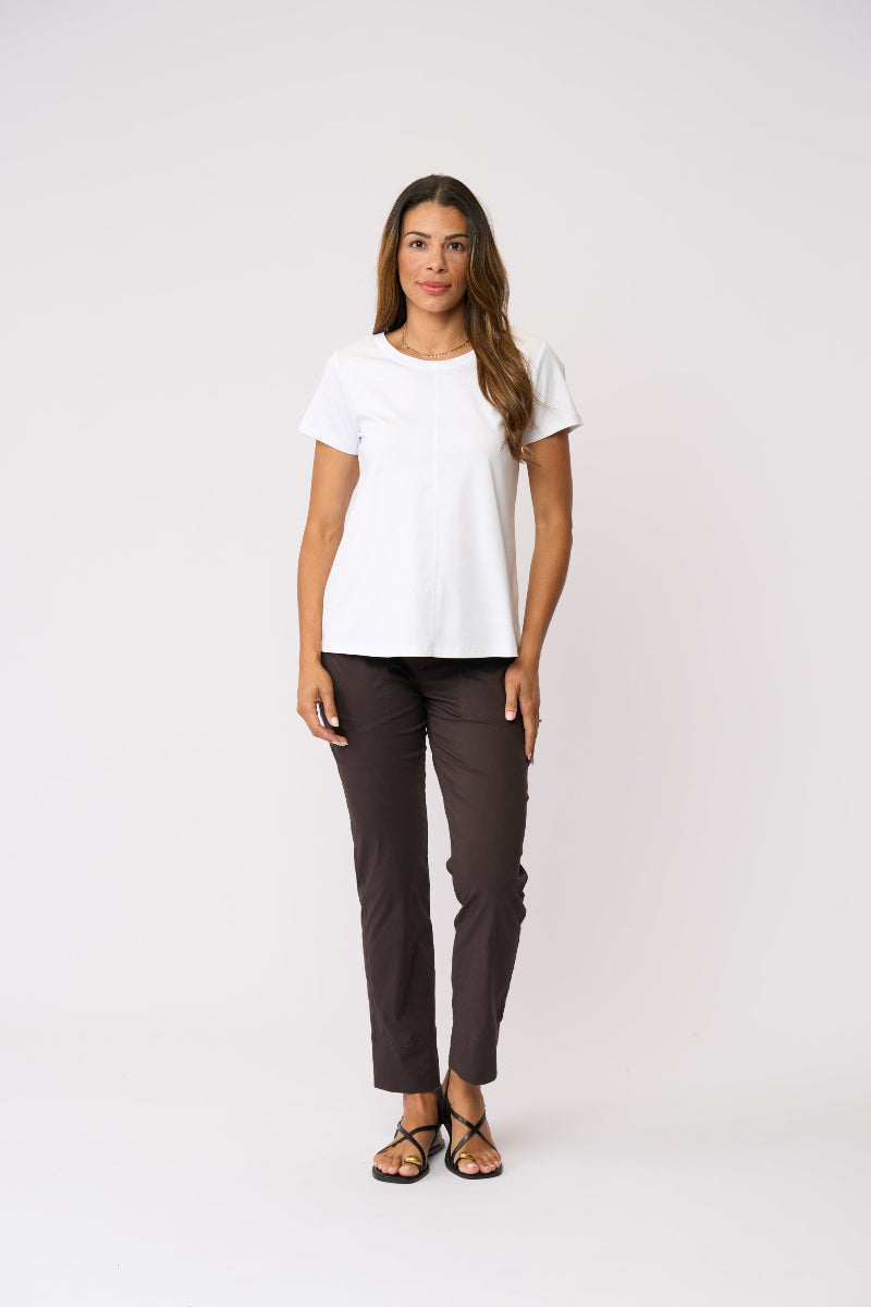 A woman modeling a white short-sleeve crewneck t-shirt and dark brown cropped pants with strappy flat sandals, standing against a light gray studio background.