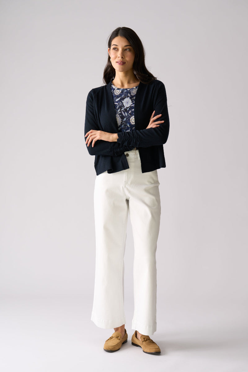 Woman wearing a black cardigan, floral top, white pants, and brown shoes on a gray background