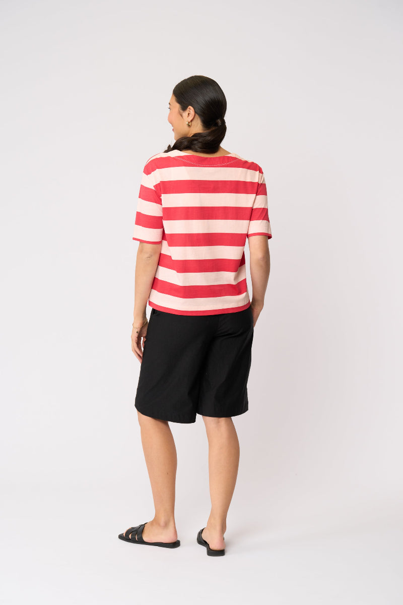 A woman is shown from behind wearing a red and white horizontally striped short-sleeve top, black shorts, and black sandals, standing against a plain light gray studio background.