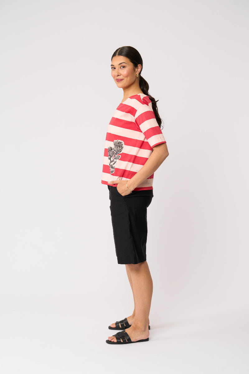 A woman stands in profile wearing a red and white striped short-sleeve top with a black rose graphic on the front, paired with black shorts and black slide sandals, against a plain light gray studio background.