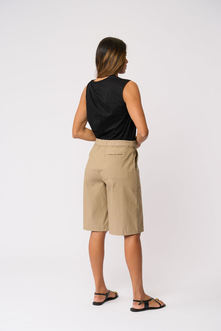 A woman shown from the back wearing a black sleeveless top and beige knee-length shorts, standing against a plain light gray studio background.