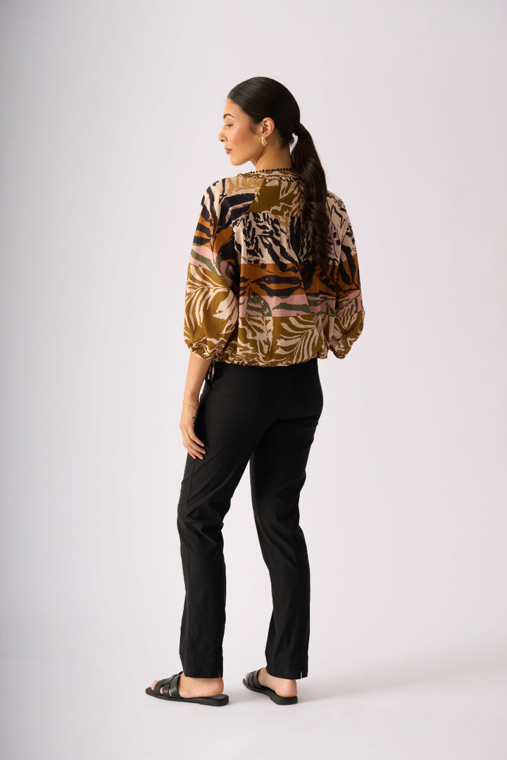 A woman shown from the back wearing a brown, black, and beige patterned blouse with voluminous sleeves, paired with black trousers and black sandals, against a light gray studio background.