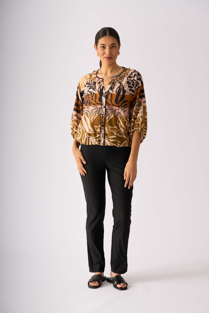 A woman modeling a three-quarter sleeve button-up blouse with an abstract botanical and animal-inspired print in brown, beige, and black, paired with black pants and black sandals, against a light gray studio background.