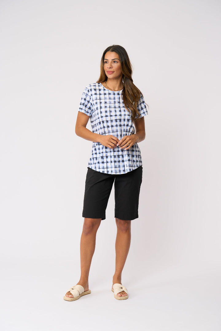 A woman modeling a short-sleeve blue and white gingham patterned top paired with black shorts and beige slide sandals, standing against a plain light gray studio background.
