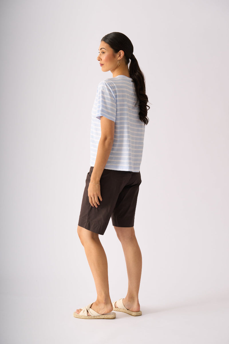 A woman shown from the side profile wearing a light blue and white striped short-sleeve t-shirt, dark brown shorts, and beige slide sandals, standing against a plain light gray studio background.