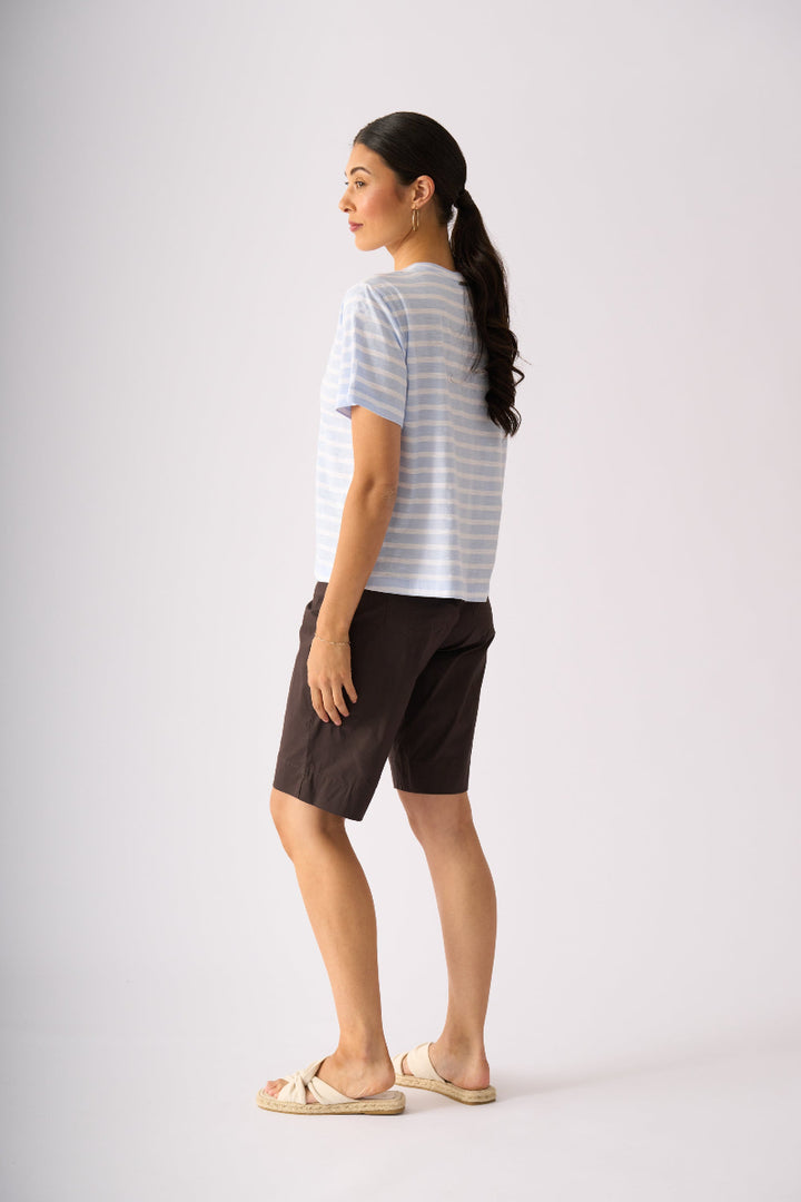 A woman shown from the side profile wearing a light blue and white striped short-sleeve t-shirt, dark brown shorts, and beige slide sandals, standing against a plain light gray studio background.