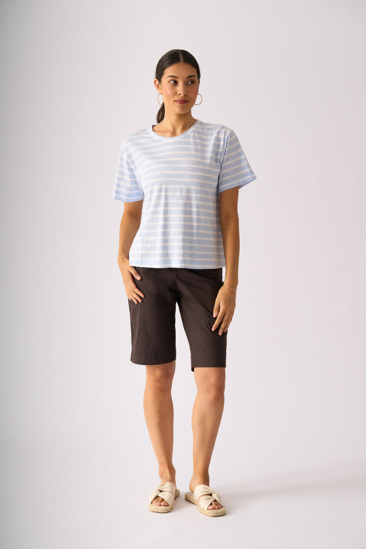A woman modeling a light blue and white striped short-sleeve t-shirt, dark brown knee-length shorts, and beige slide sandals against a light gray studio background.