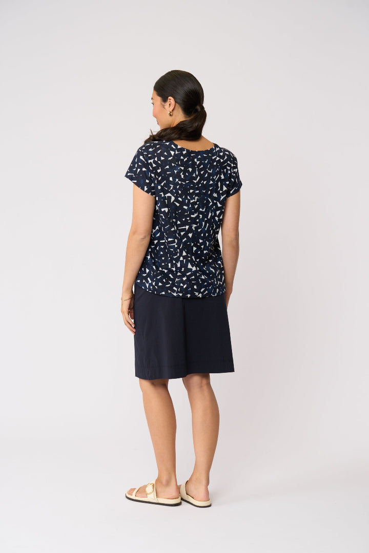 A woman shown from the back wearing a navy blue short-sleeve top with a white abstract pattern and a matching navy blue skirt, standing against a light gray studio background.