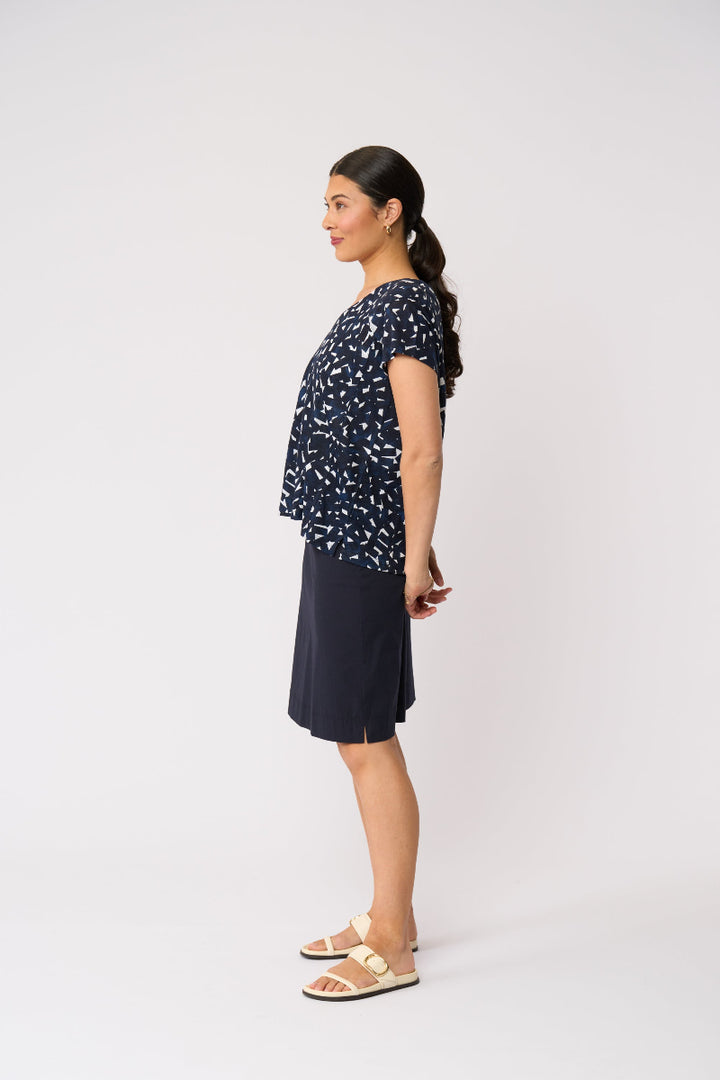 A woman stands in profile wearing a navy short-sleeve top with a small white abstract print and a navy skirt, paired with beige slide sandals, against a plain light gray studio background.