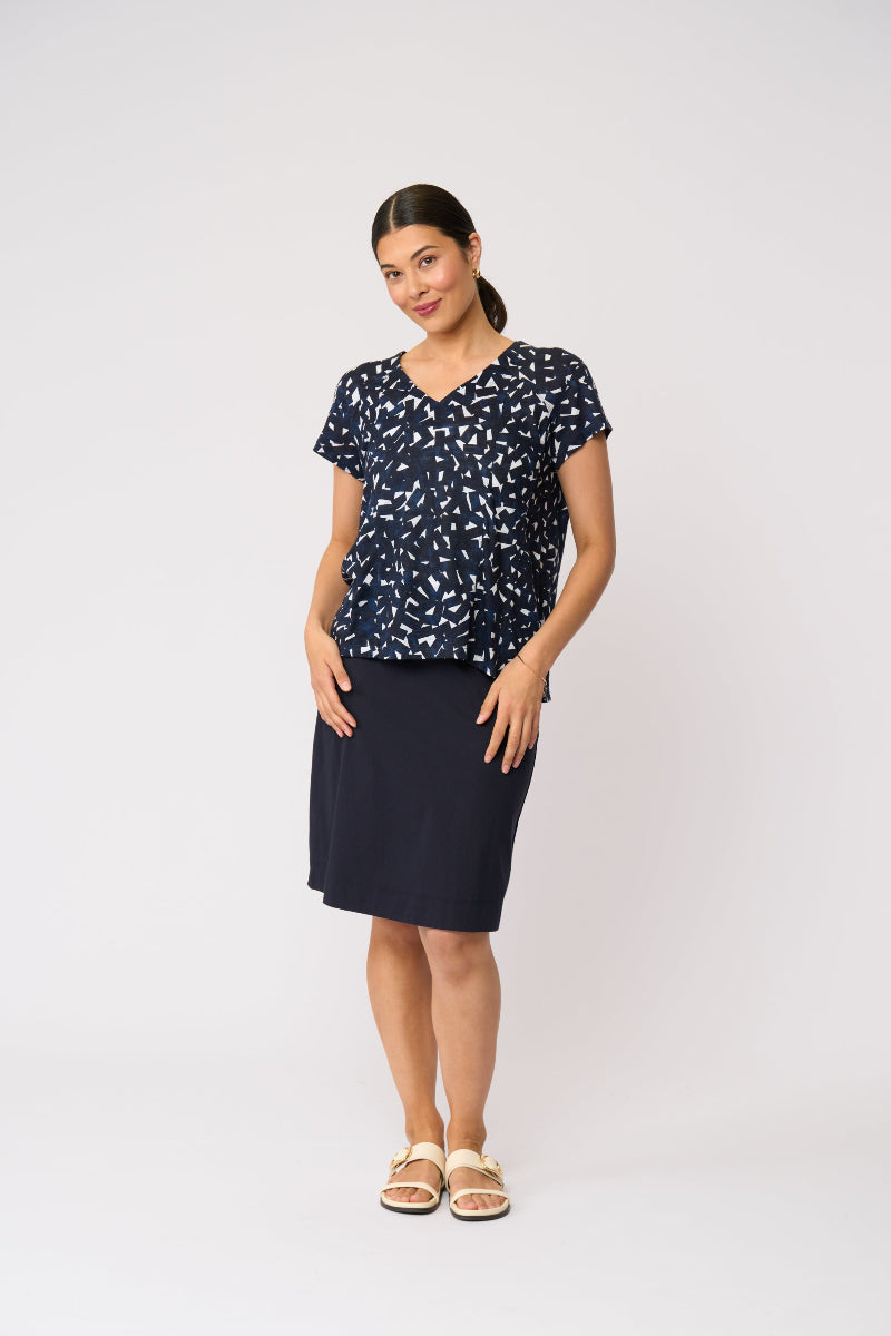 A woman modeling a two-piece navy blue set consisting of a short-sleeve V-neck top with a white abstract pattern and a solid navy blue knee-length skirt, paired with beige slide sandals, against a light gray studio background.