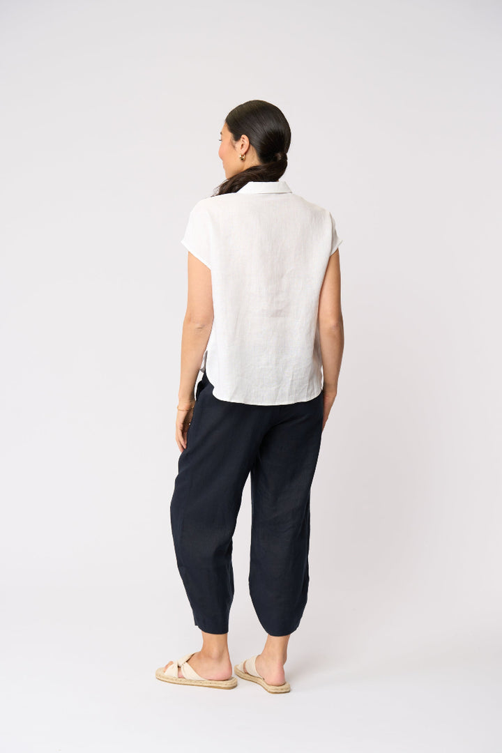 A woman is shown from behind wearing a white short-sleeve linen shirt and dark navy wide-leg linen pants, paired with beige slide sandals, against a plain white studio background.