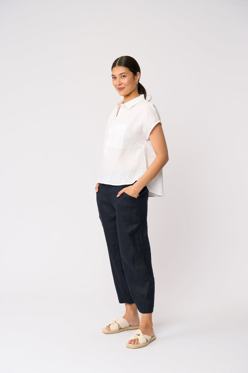 A woman modeling a white short-sleeve collared top and dark navy cropped pants with beige slide sandals, standing against a plain light gray studio background.