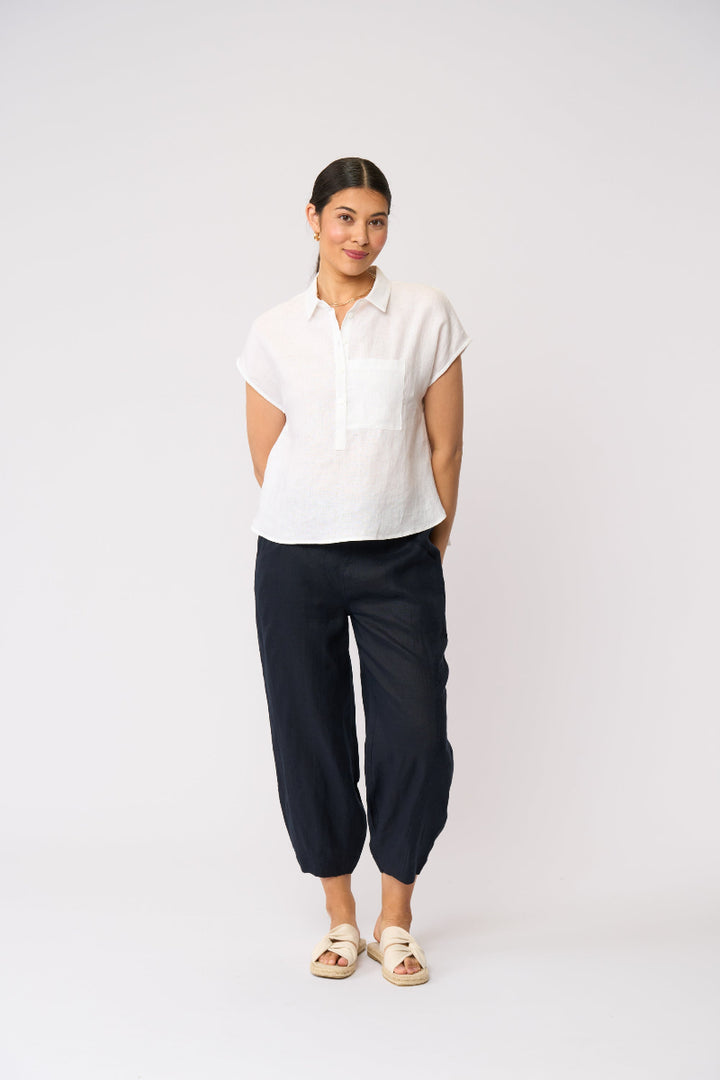 A woman modeling a white short-sleeve button-up shirt and dark navy cropped pants, standing against a plain light gray studio background.