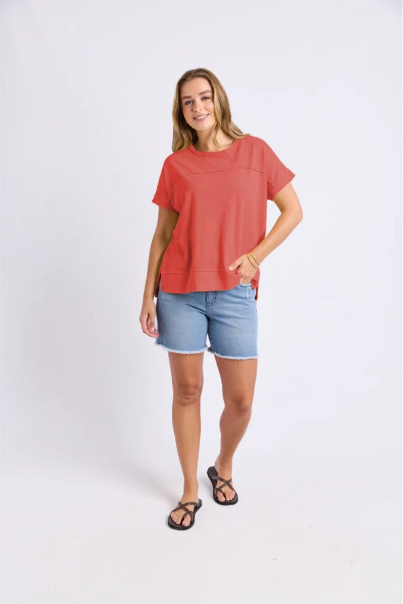 A woman modeling a coral-red short-sleeve crewneck top and light-wash denim shorts against a plain light gray studio background.