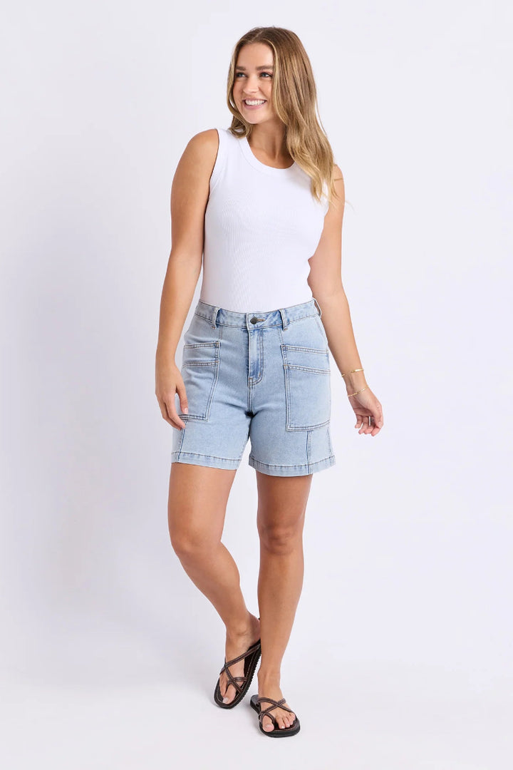A woman modeling light-wash denim shorts with a white sleeveless top and black sandals, standing against a plain white studio background.