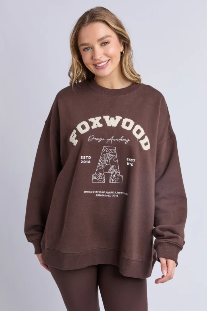Woman wearing a brown sweatshirt with 'FOXWOOD' printed on it against a plain background