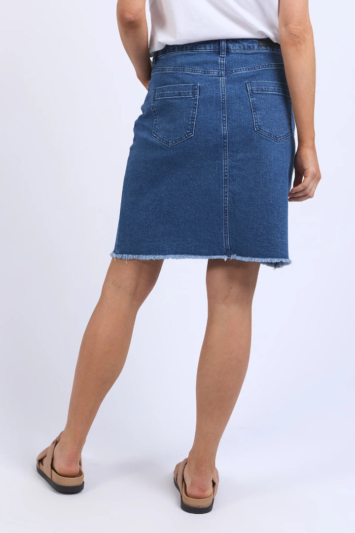 A person shown from the back wearing a blue denim skirt with a frayed hem, paired with a white top and tan slide sandals, against a plain light gray studio background.