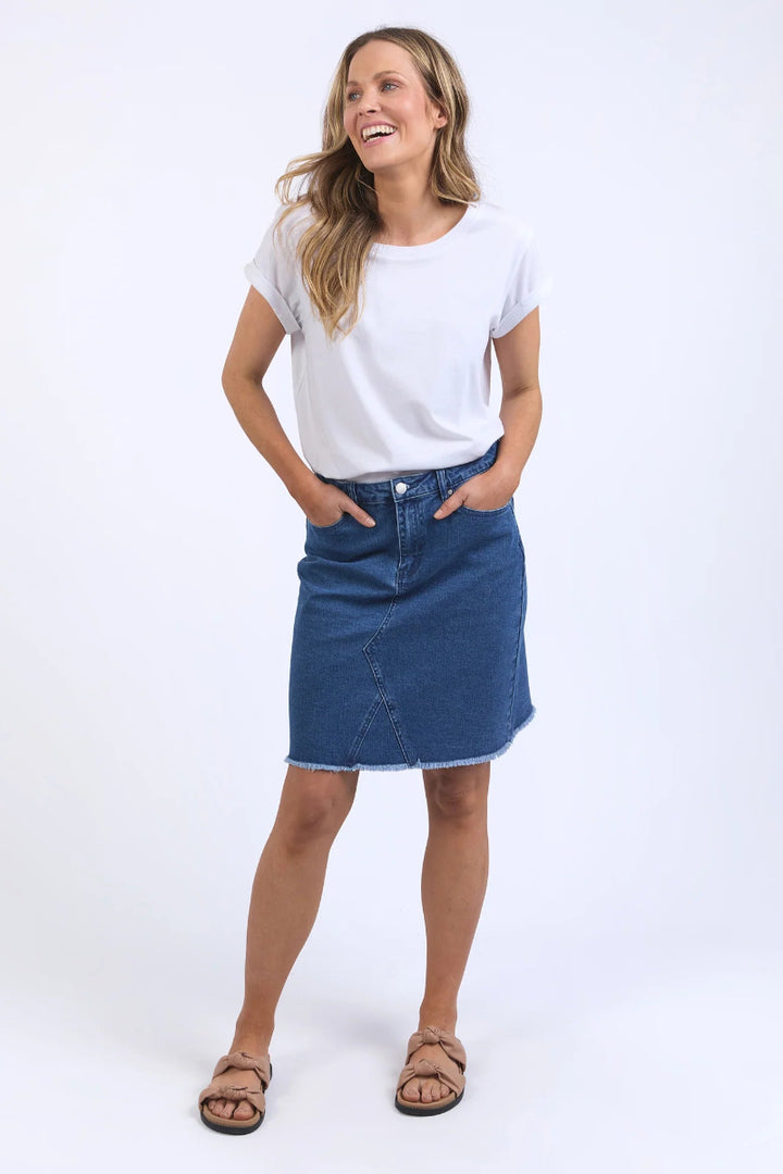 A woman modeling a blue denim mini skirt with a white t-shirt and brown sandals, standing against a plain white studio background.