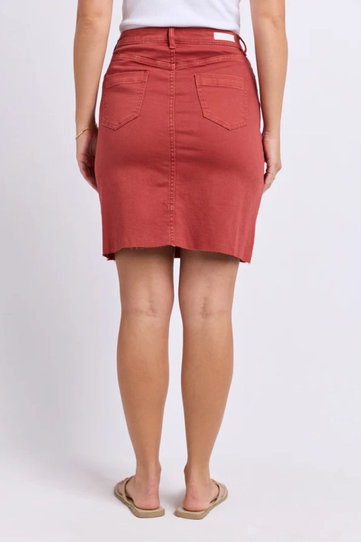 A woman shown from the back wearing a rust-red denim mini skirt with a button closure, belt loop, and two back patch pockets, paired with a white top and tan sandals against a light gray studio background.