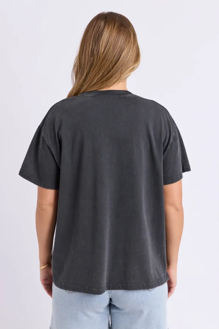 A woman viewed from the back wearing a faded black washed short-sleeve t-shirt and light blue jeans, set against a plain light gray studio background.