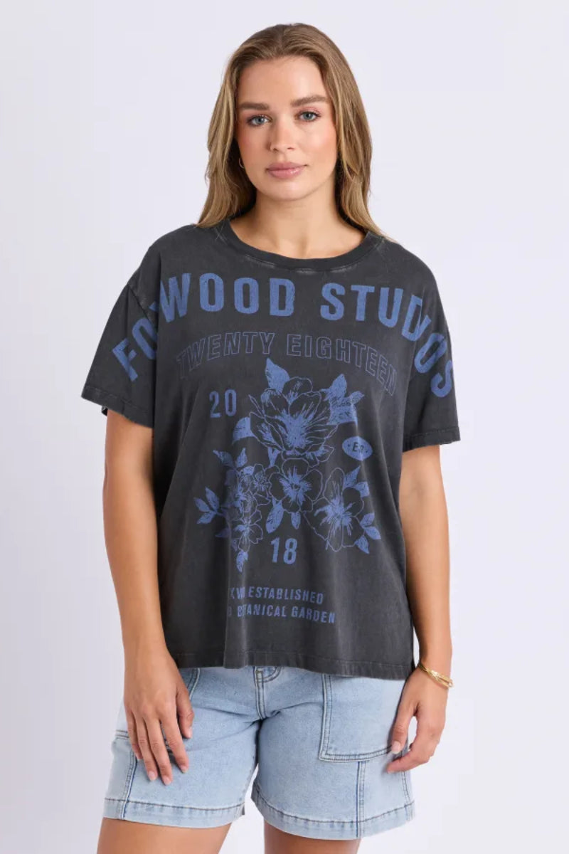 A woman modeling a dark gray graphic t-shirt with blue text and floral designs reading 'FOWOOD STUDIOS TWENTY EIGHTER', paired with light blue denim shorts, against a light gray studio background.