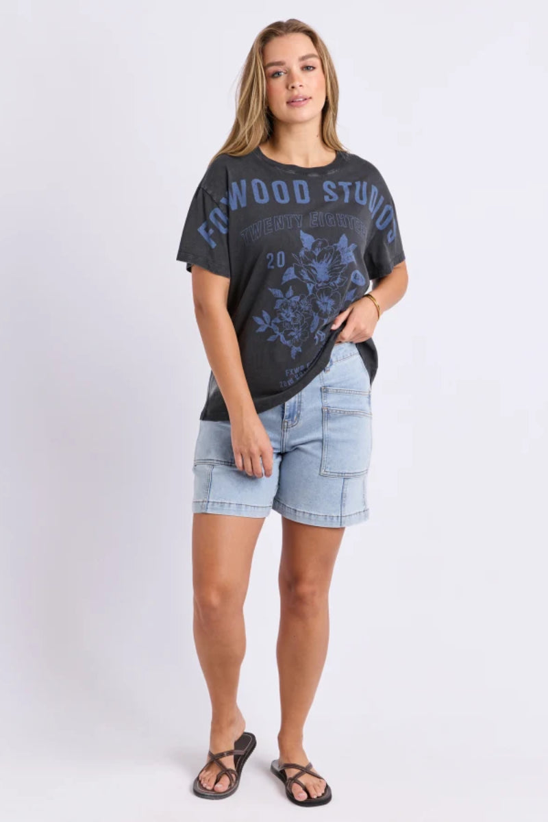 A woman modeling a dark charcoal gray graphic t-shirt with blue text and floral print, paired with light wash denim shorts and black sandals, against a light gray studio background.