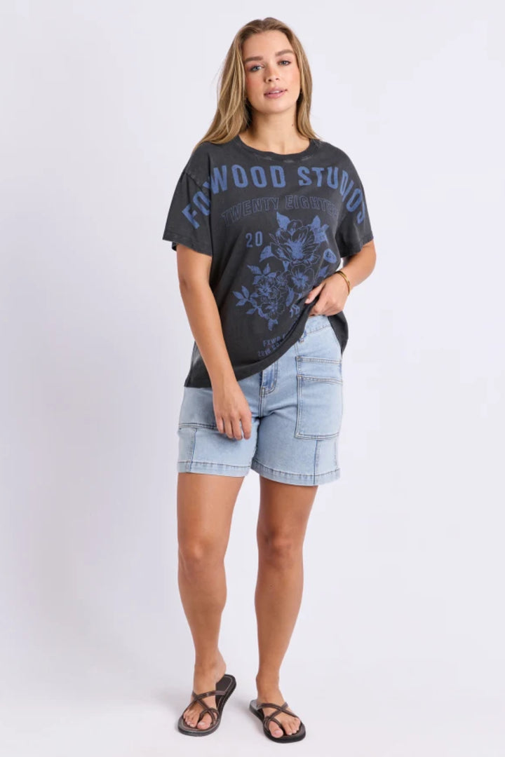 A woman modeling a dark charcoal gray graphic t-shirt with blue text and floral print, paired with light wash denim shorts and black sandals, against a light gray studio background.