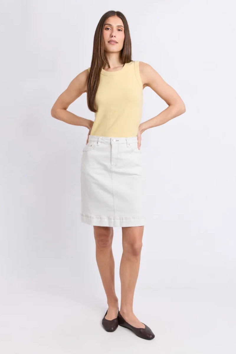 A woman modeling a pale yellow sleeveless knit top and a white denim skirt, paired with dark brown flats, against a light gray studio background.