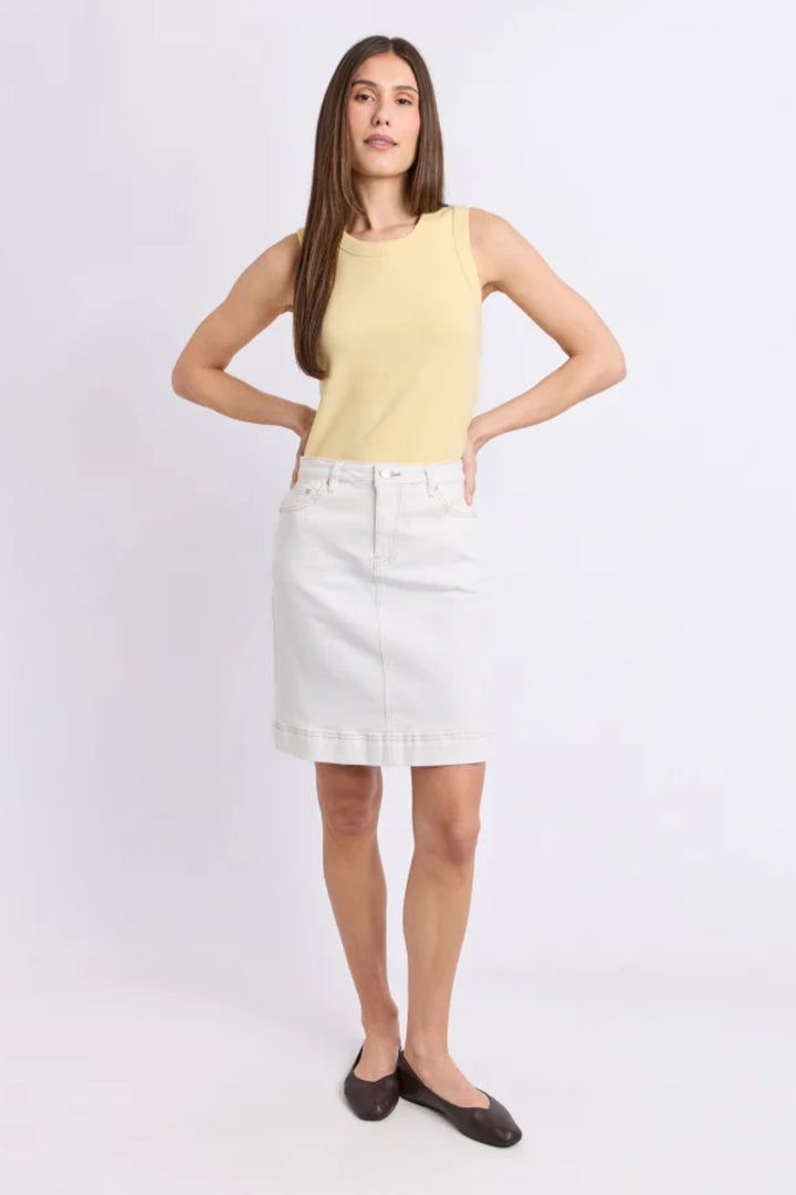 A woman modeling a pale yellow sleeveless knit top and a white denim skirt, paired with dark brown flats, against a light gray studio background.