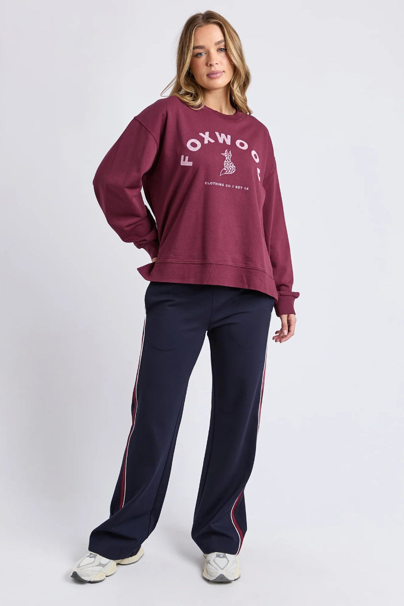 Woman wearing a maroon sweatshirt with 'Foxwood' logo and navy tracksuit pants on a white background