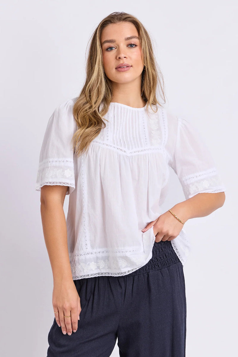 A woman modeling a white short-sleeve blouse with delicate lace trim and a gathered front, paired with dark navy pants, against a light gray studio background.