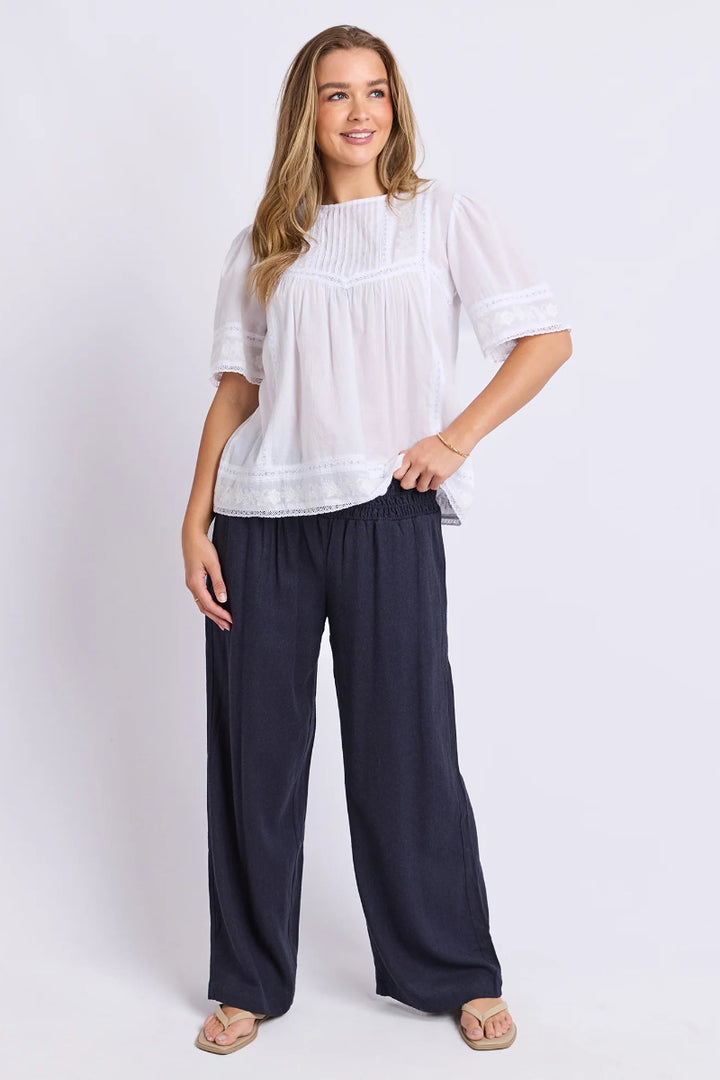 A woman modeling a white short-sleeve blouse with delicate lace trim and dark navy wide-leg pants, standing against a light gray studio background.