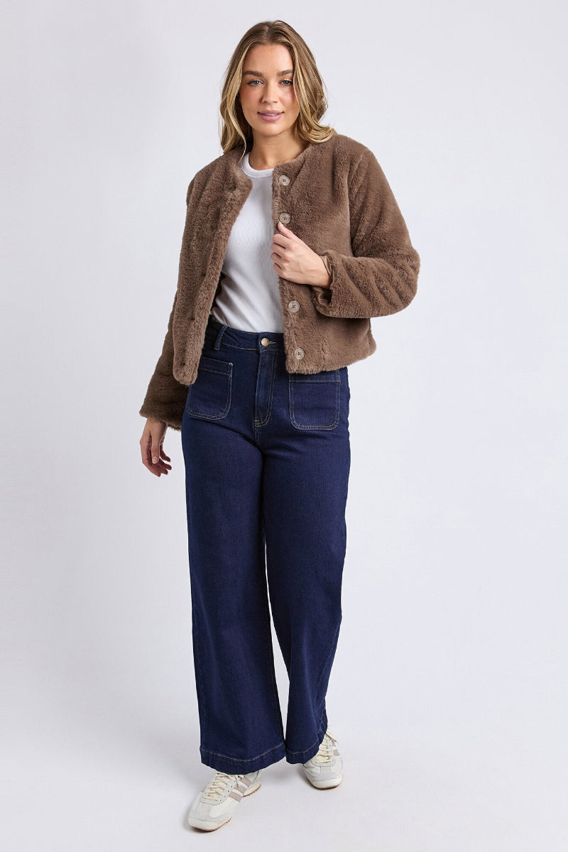 Woman wearing a brown jacket, white shirt, blue jeans, and white sneakers on a light gray background