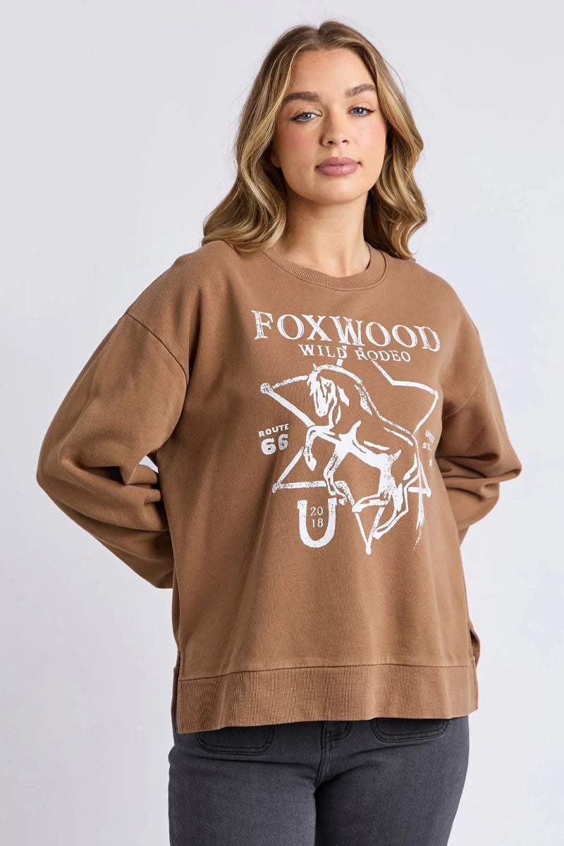 Woman wearing a brown sweatshirt with 'Foxwood Wild Rodeo' print on a plain background