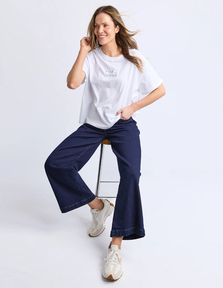 Woman seated on stool wearing white Foxwood T-shirt, dark jeans, and sneakers.