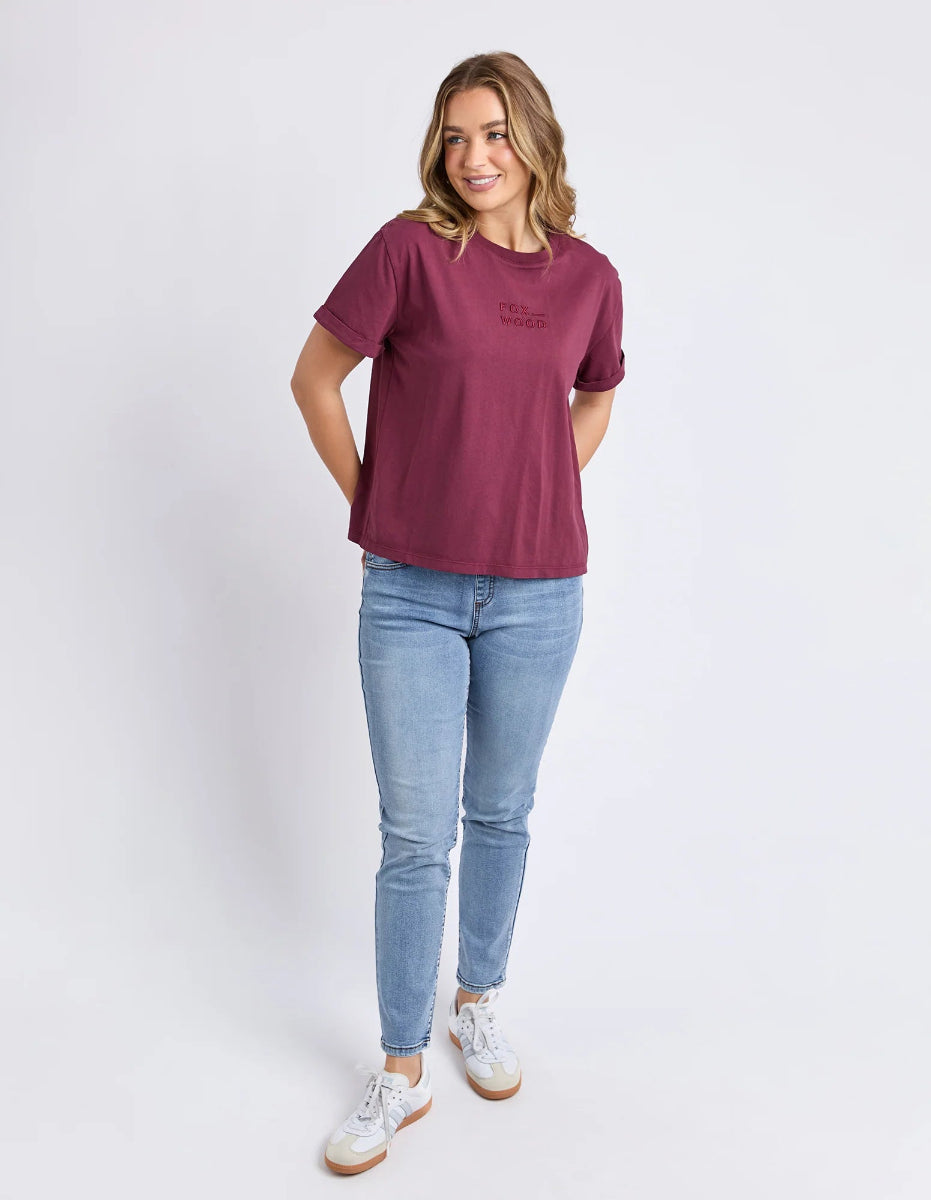 Woman in wine Foxwood T-shirt, light blue jeans, and white sneakers standing.