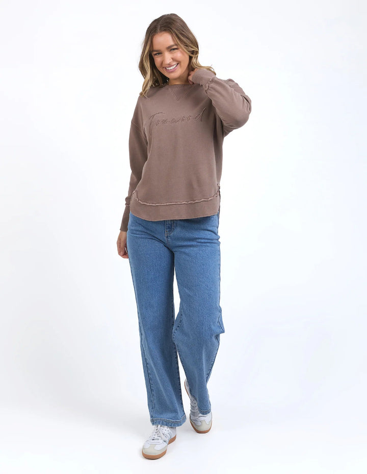 Woman wearing brown Foxwood sweatshirt, blue jeans, and white sneakers.