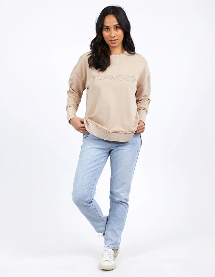 Woman in beige Foxwood sweatshirt, light blue jeans, and white sneakers standing.