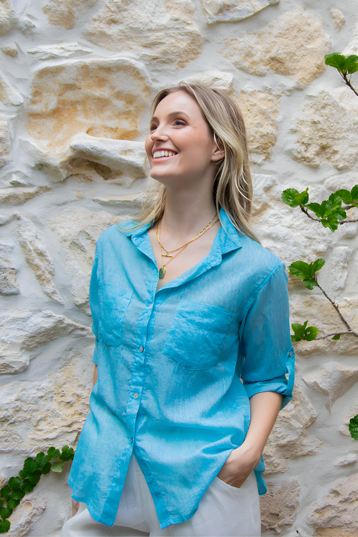 Woman wearing a blue shirt button shirt standing against a stone wall with greenery.