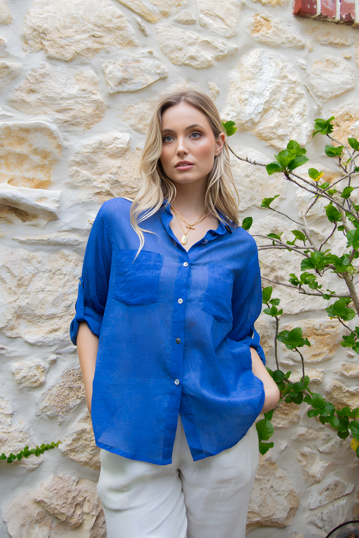 Woman wearing a blue shirt and white pants standing against a stone wall with greenery.