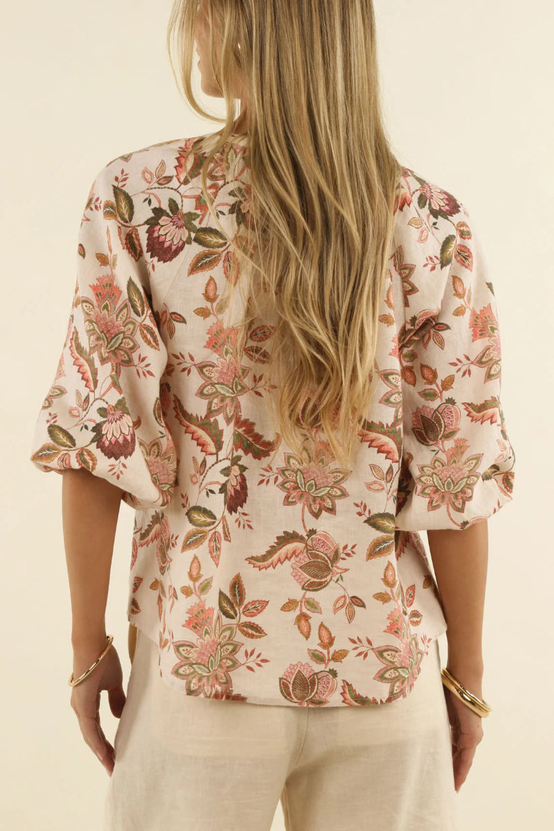 A woman viewed from the back wearing a cream-colored blouse with a pink, brown, and green floral pattern and three-quarter sleeves, paired with light beige pants against a warm off-white background.