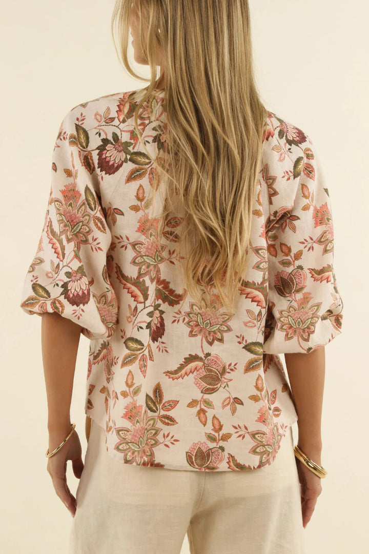 A woman viewed from the back wearing a cream-colored blouse with a pink, brown, and green floral pattern and three-quarter sleeves, paired with light beige pants against a warm off-white background.