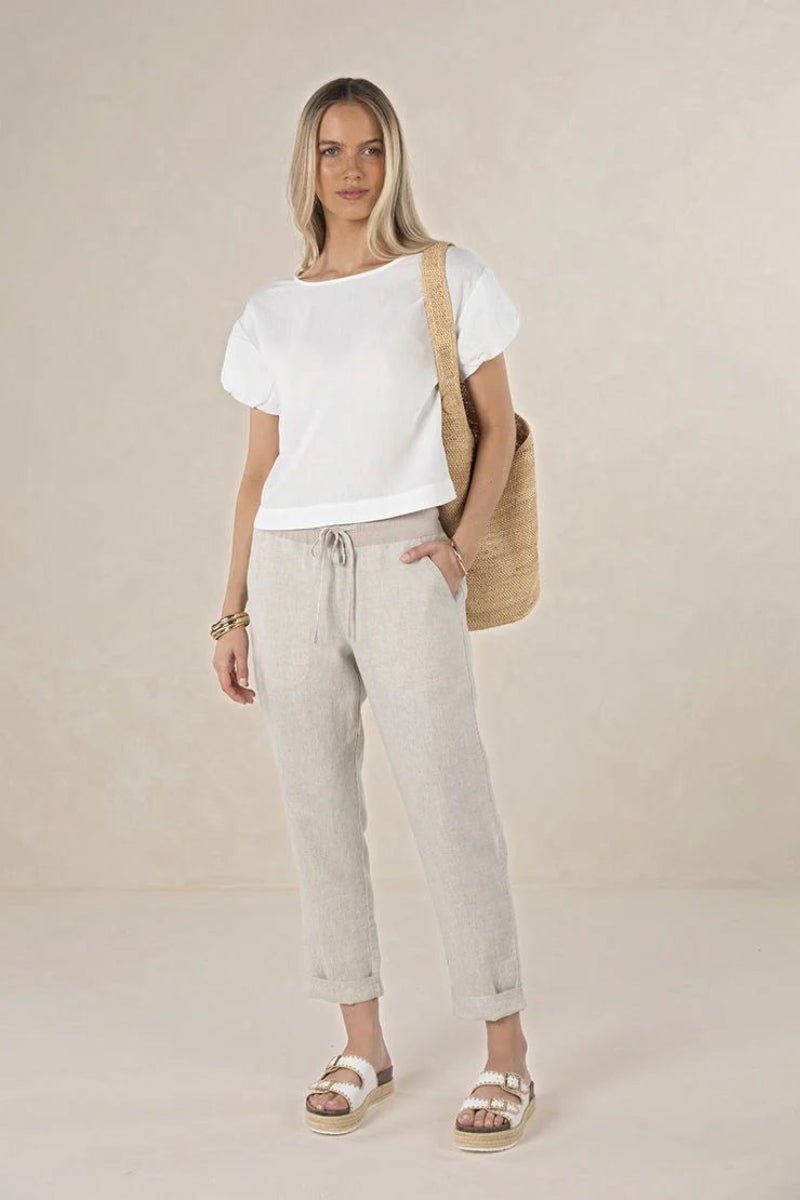 A woman modeling a white short-sleeve t-shirt and light beige linen drawstring pants, styled with a beige woven shoulder bag and white platform sandals against a neutral beige studio backdrop.