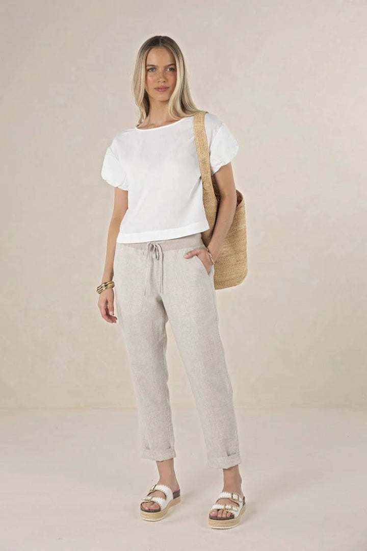 A woman modeling a white short-sleeve t-shirt and light beige linen drawstring pants, styled with a beige woven shoulder bag and white platform sandals against a neutral beige studio backdrop.