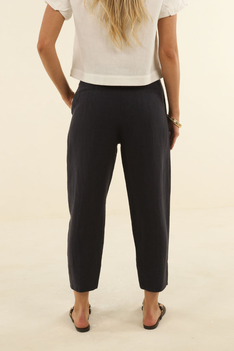 A woman shown from the back wearing black cropped trousers, a white short-sleeve top, and black sandals against a plain cream studio background.
